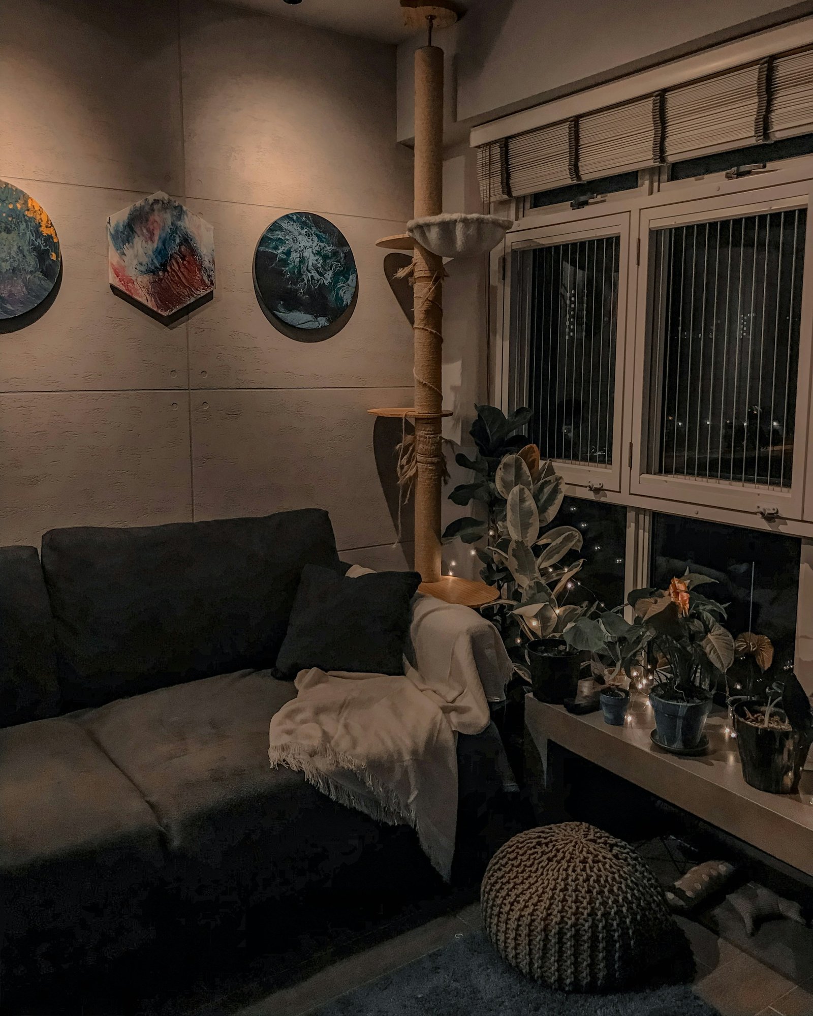 A cozy living room with layered lamps, textured cushions, a knitted throw, indoor plants, books, and personal artwork.